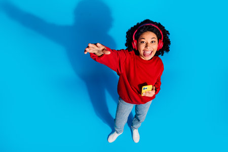Full length photo of lovely young lady top view stretch arm headphones device dressed stylish red garment isolated on blue color backgroundの写真素材