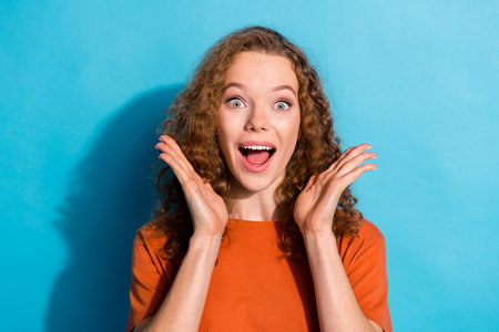Photo portrait of youth beautiful funny girl with wavy hair wear orange t shirt raised hands up surprised isolated on blue color backgroundの写真素材