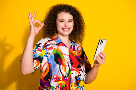 Photo portrait of pretty young girl hold device show okey wear trendy colorful print outfit isolated on yellow color backgroundの写真素材