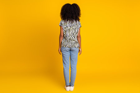Rear view photo of casual young charming woman curly hair in trendy zebra print shirt and jeans isolated on yellow color backgroundの写真素材