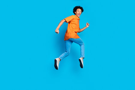 Full length photo of attractive funny guy wear orange shirt running jumping high emtpy space isolated blue color backgroundの写真素材