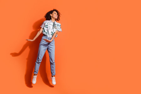 Full length photo of american woman in jeans and hawaiian shirt jumping dreaming looking excited isolated on orange color backgroundの写真素材