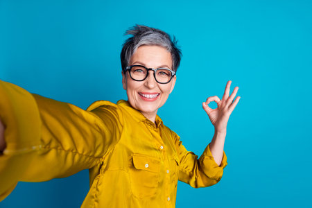 Photo of adorable good mood lady dressed shirt eyewear tacking selfie showing okey isolated blue...