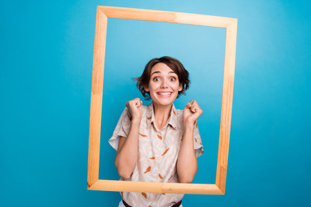 Portrait of delighted positive girl wooden photo frame raise fists success isolated on blue color...