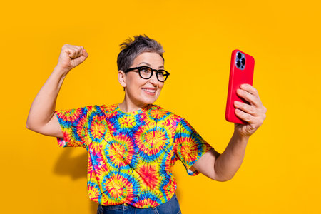 Portrait of nice aged lady use phone raise fist wear t-shirt isolated on yellow color background