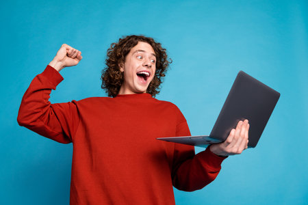 Photo of handsome excited guy wear red sweater winning game modern device isolated blue color...