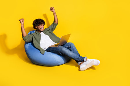 Full body length photo of successful guy lying pouf chilling playing computer raised fists up isolated on yellow color backgroundの写真素材