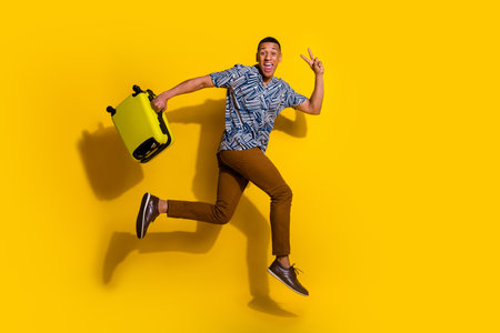 Full length photo of cheerful positive guy dressed print shirt jumping running holding luggage empty space isolated yellow color backgroundの写真素材