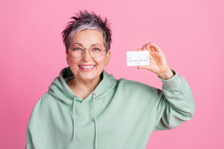 Photo of cheerful cool lady dressed khaki sweatshirt showing credit card empty space isolated pink...