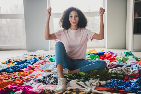 Full size photo of nice young woman sit messy pile stack clothes raise fists apartment indoors