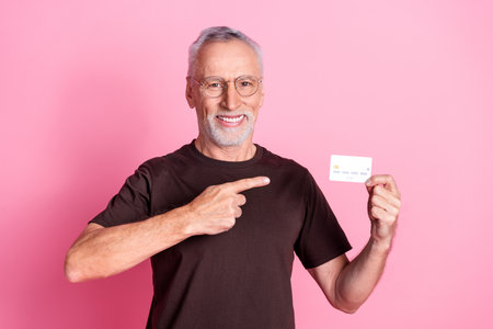 Portrait of optimistic senior man with beard dressed t-shirt in glasses indicating at credit card...