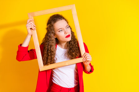 Portrait of gorgeous young lady wooden frame look empty space wear red suit isolated on yellow color backgroundの写真素材