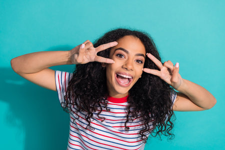 Photo portrait of lovely young lady show v-sign posing dressed stylish striped garment isolated on cyan color backgroundの写真素材