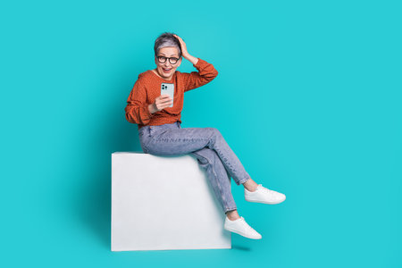 Full body photo of attractive senior woman sit white cube amazed hold device dressed brown clothes isolated on cyan color backgroundの写真素材