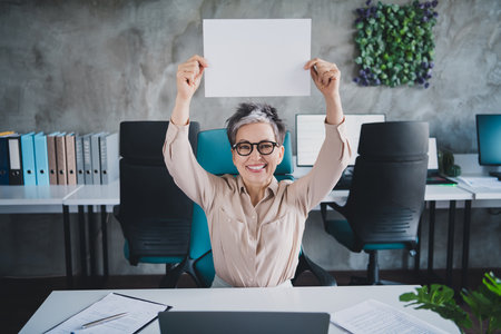 Photo of attractive good mood age lady agent dressed formal shirt rising paper empty space indoors workplace workstation loftの写真素材