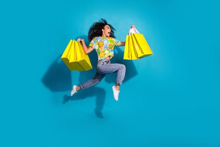 Full length photo of adorable lovely girl wear flower print t-shirt jumping holding shoppers empty space isolated blue color backgroundの写真素材