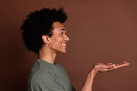 Side photo of young positive guy with curly haircut wearing gray t shirt smiling holding two arms asking help isolated on brown color backgroundの写真素材