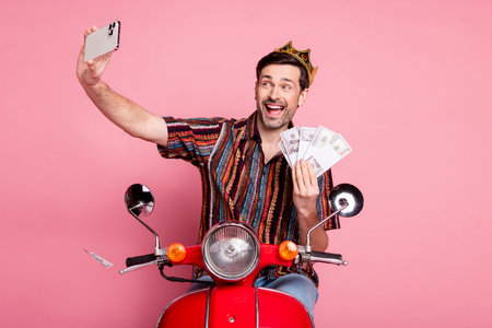 Portrait of young nice man wear shirt crown drive bike phone make selfie money isolated on pink...