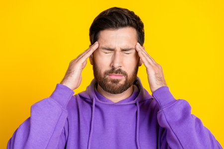 Portrait of tired suffering man with stylish bristle dressed purple hoodie eyes closed have headache isolated on yellow color backgroundの写真素材