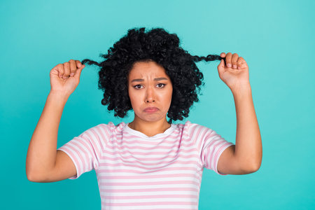 Photo of young upset girl in striped t shirt touching her curls hate bad quality hair shampoo isolated on aquamarine color backgroundの写真素材