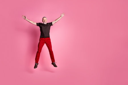 Full size photo of overjoyed senior man dressed brown t-shirt red trousers jumping near empty space isolated on pink color backgroundの写真素材