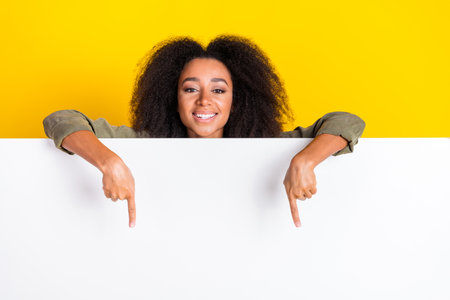Photo of cheerful girl with perming coiffure wear khaki shirt directing on promo on board empty space isolated on yellow color backgroundの写真素材