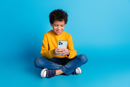 Full body photo of cute little boy sit floor hold device dressed stylish yellow clothes isolated on blue color backgroundの写真素材