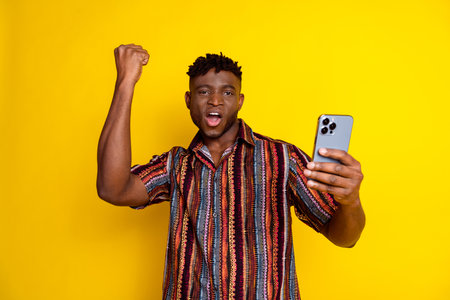 Portrait of nice young man smart phone raise fist wear shirt isolated on yellow color backgroundの写真素材