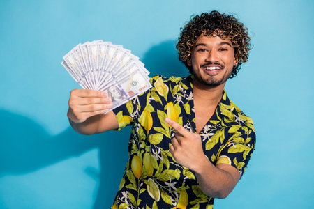 Photo of overjoyed guy with beard wavy hairstyle dressed print shirt indicating at dollars in arm...