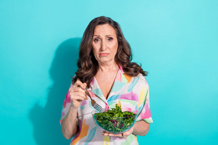 Photo portrait of attractive senior woman unhappy eat salad dressed stylish colorful clothes isolated on cyan color backgroundの写真素材