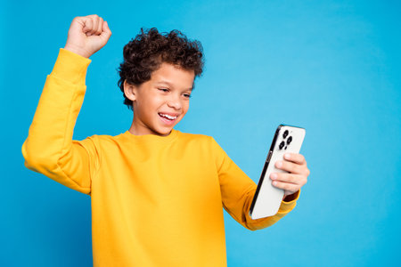Photo portrait of cute little boy winning hold device dressed stylish yellow clothes isolated on blue color backgroundの写真素材