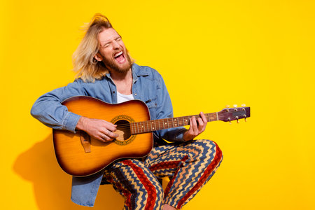 Photo portrait of handsome young male play guitar musician singing dressed stylish denim garment isolated on yellow color backgroundの写真素材