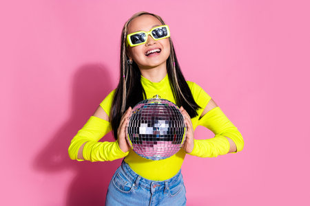 Photo of positive asian girl with dyed hairstyle dressed yellow shirt in sunglass holding disco ball isolated on pink color backgroundの写真素材