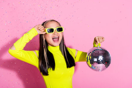 Photo of ecstatic cool girl with dyed hairstyle dressed yellow turtleneck touch sunglass hold discoball isolated on pink color backgroundの写真素材