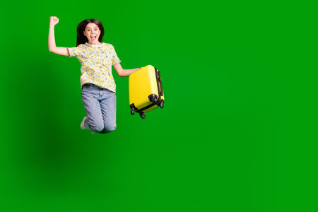Full length photo of lovely teen lady jump winning hold suitcase dressed stylish flowers print garment isolated on green color backgroundの写真素材