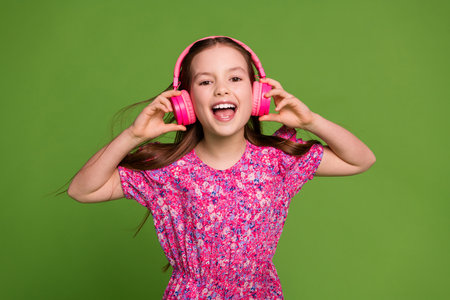 Portrait of lovely schoolgirl touch headphones wear pink dress isolated on green color background
