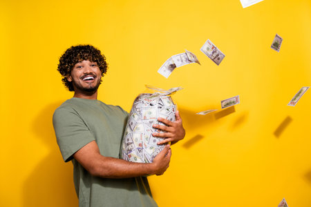 Photo of optimistic satisfied guy with wavy hair dressed khaki t-shirt holding jar with flying dollars isolated on yellow color backgroundの写真素材