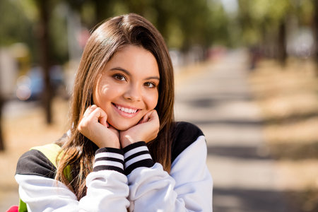 Photo of adorable optimistic student with straight hairstyle dressed stylish jacket hold hands under chin in sunny weather outdoorsの写真素材