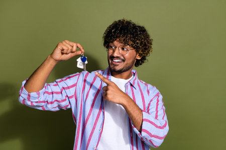 Photo of overjoyed guy with stubble wavy hairstyle dressed striped shirt directing look at keys in arm isolated on khaki color backgroundの写真素材