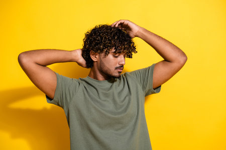 Photo of cool handsome guy with wavy hair dressed t-shirt holding arms on head after barber shop isolated on yellow color backgroundの写真素材