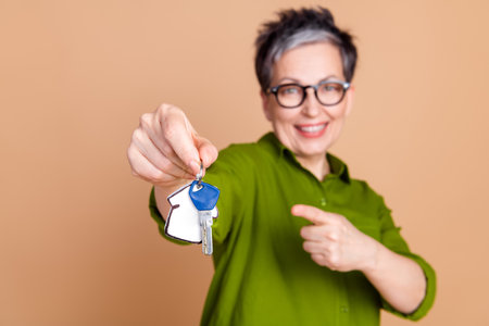 Photo portrait of pretty senior female hold point house keys wear trendy green outfit isolated on beige color backgroundの写真素材