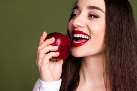 Cropped photo of pretty young girl eating apple wear trendy white outfit isolated on khaki color backgroundの写真素材