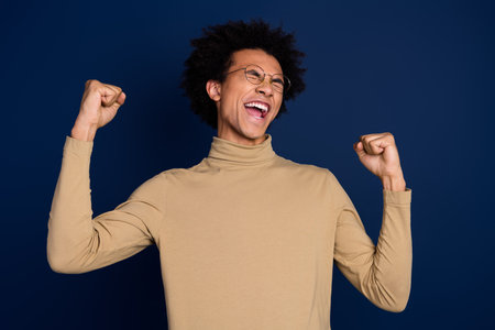 Photo portrait of handsome young man raise fists winning dressed stylish beige garment isolated on dark blue color backgroundの写真素材