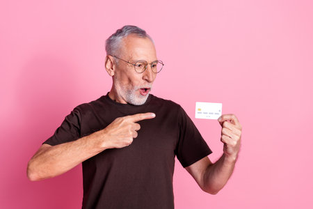 Portrait of impressed senior man with beard dressed t-shirt in glasses indicating look at credit card isolated on pink color backgroundの写真素材
