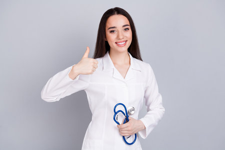 Portrait of qualified doctor woman show thumb up white coat isolated on grey color backgroundの写真素材
