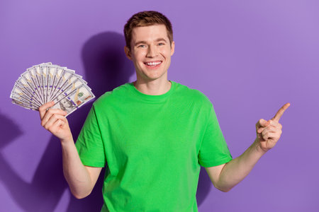 Portrait of nice young man hold cash point finger empty space wear t-shirt isolated on purple color backgroundの写真素材