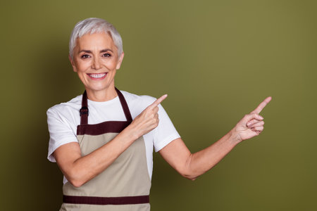 Photo of adorable happy woman wear apron serving beverages pointing two fingers empty space isolated khaki color backgroundの写真素材