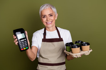 Photo of adorable happy woman wear apron serving beverages holding terminal empty space isolated khaki color backgroundの写真素材