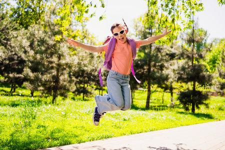 Full body photo of positive happy little girl smile summer jumping sunglass backpack good mood weekend enjoy outdoors outside park streetの写真素材