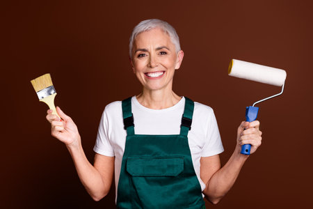 Photo of cheerful charming senior woman wear green workwear hold equipment isolated on brown color backgroundの写真素材